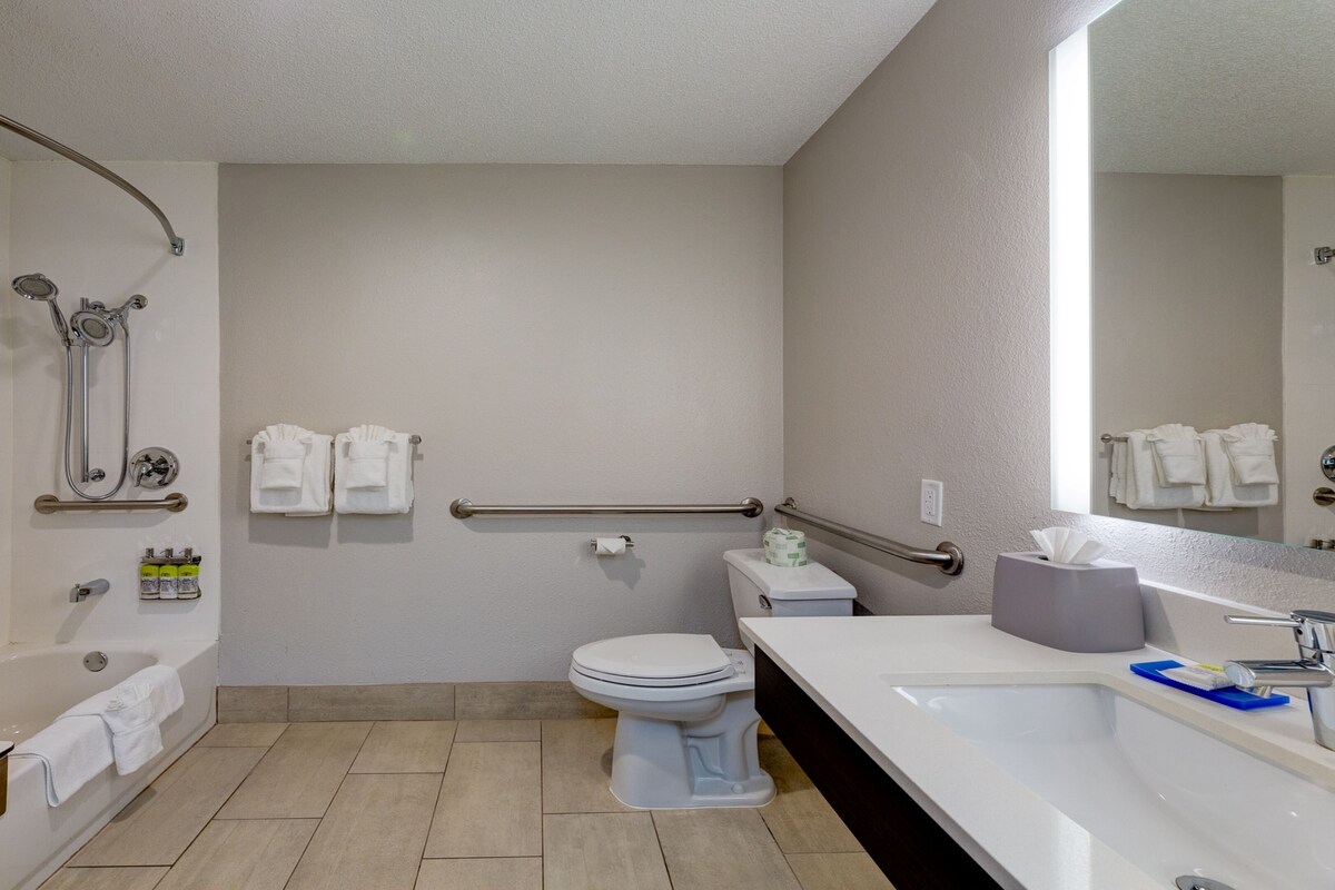 A modern bathroom with a bathtub, toilet, and sink. There are handrails and a showerhead.