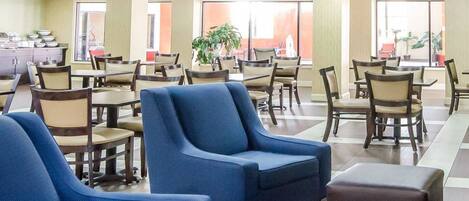 A spacious dining area with multiple tables and chairs, a kitchenette in the background, and large windows allowing natural light.