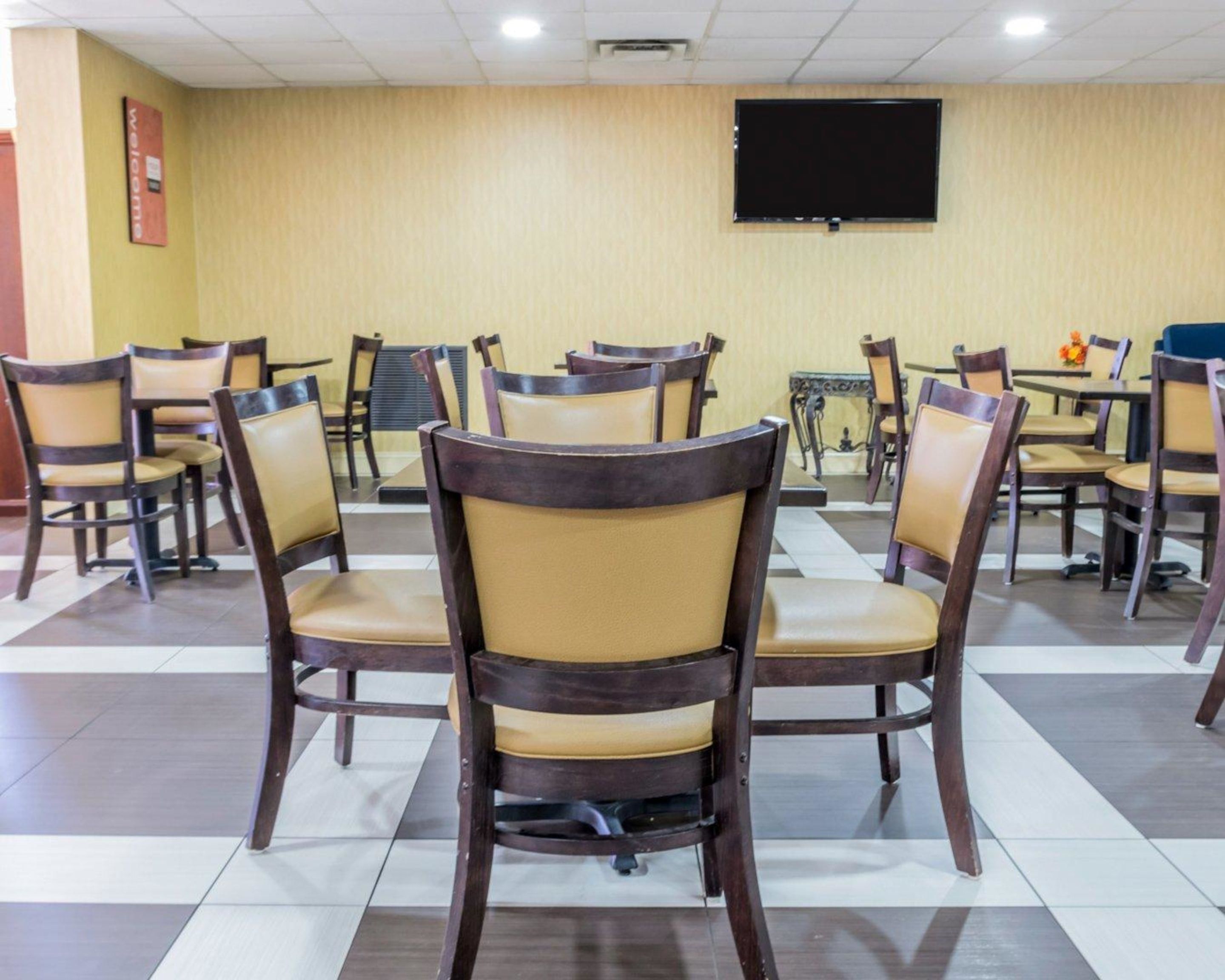 A hotel room with a checkered floor, wooden chairs with cushioned seats, a flat-screen TV mounted on the wall, and a 'Welcome' sign on the wall.