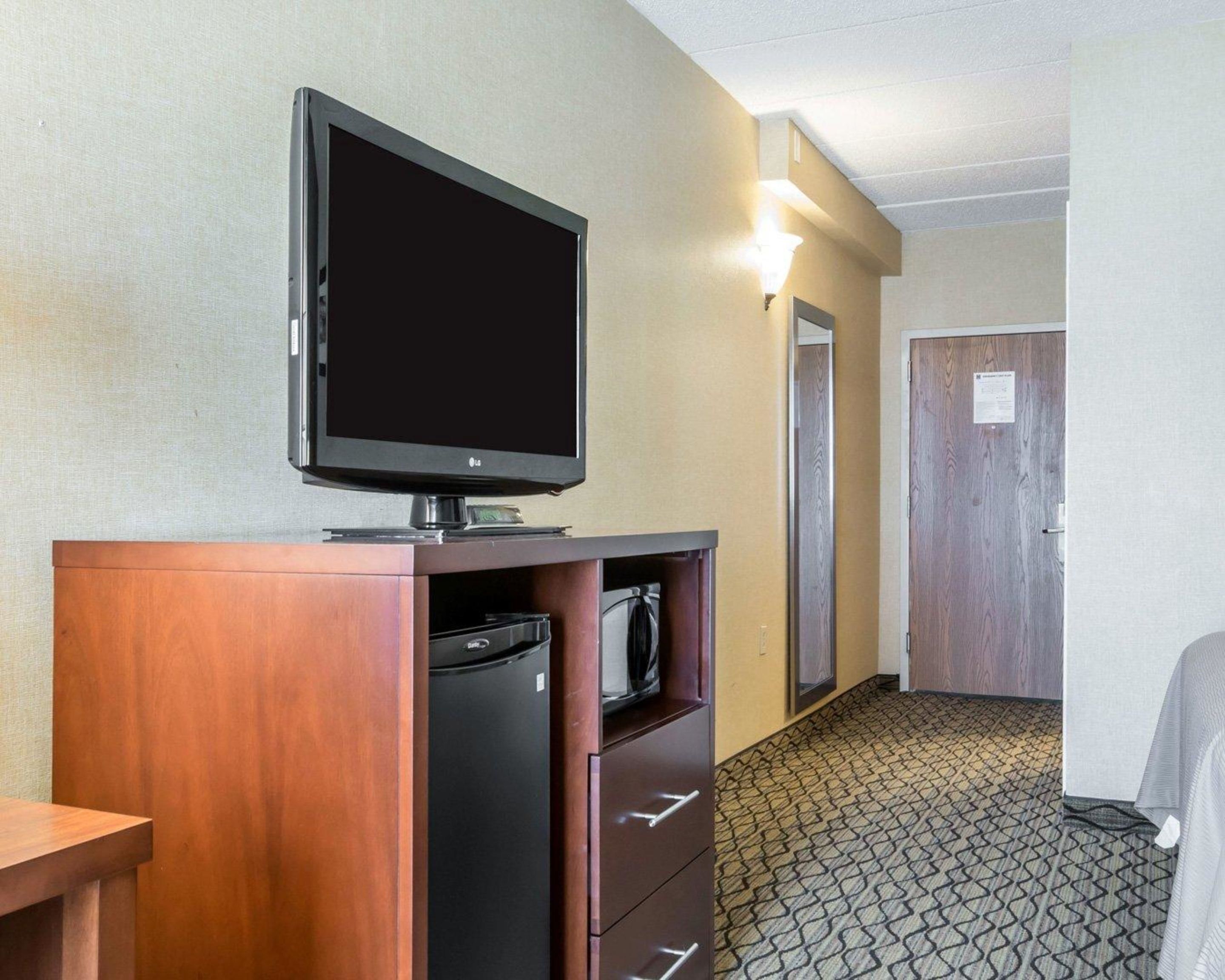 Une chambre d’hôtel équipée d’un téléviseur à écran plat posé sur un meuble en bois, d’un mini-réfrigérateur et d’un four à micro-ondes. On y trouve une porte avec une lumière au-dessus et un tapis à motifs.