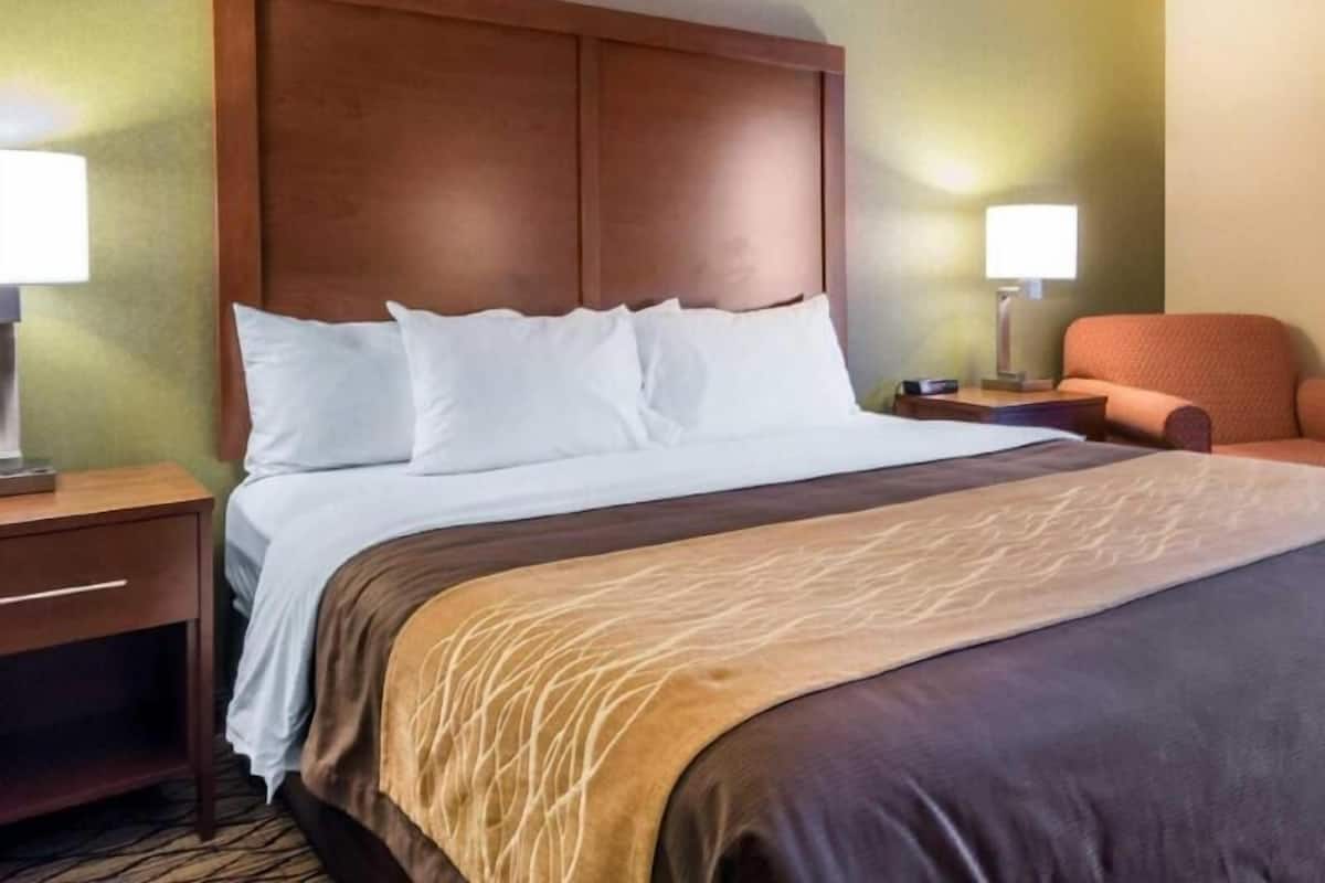 A hotel room with a large bed, a wooden headboard, a nightstand with a lamp, and a chair.