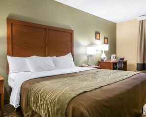A hotel room with a large bed, a wooden headboard, a bedside table with a lamp, a desk with a chair, and a window with curtains.