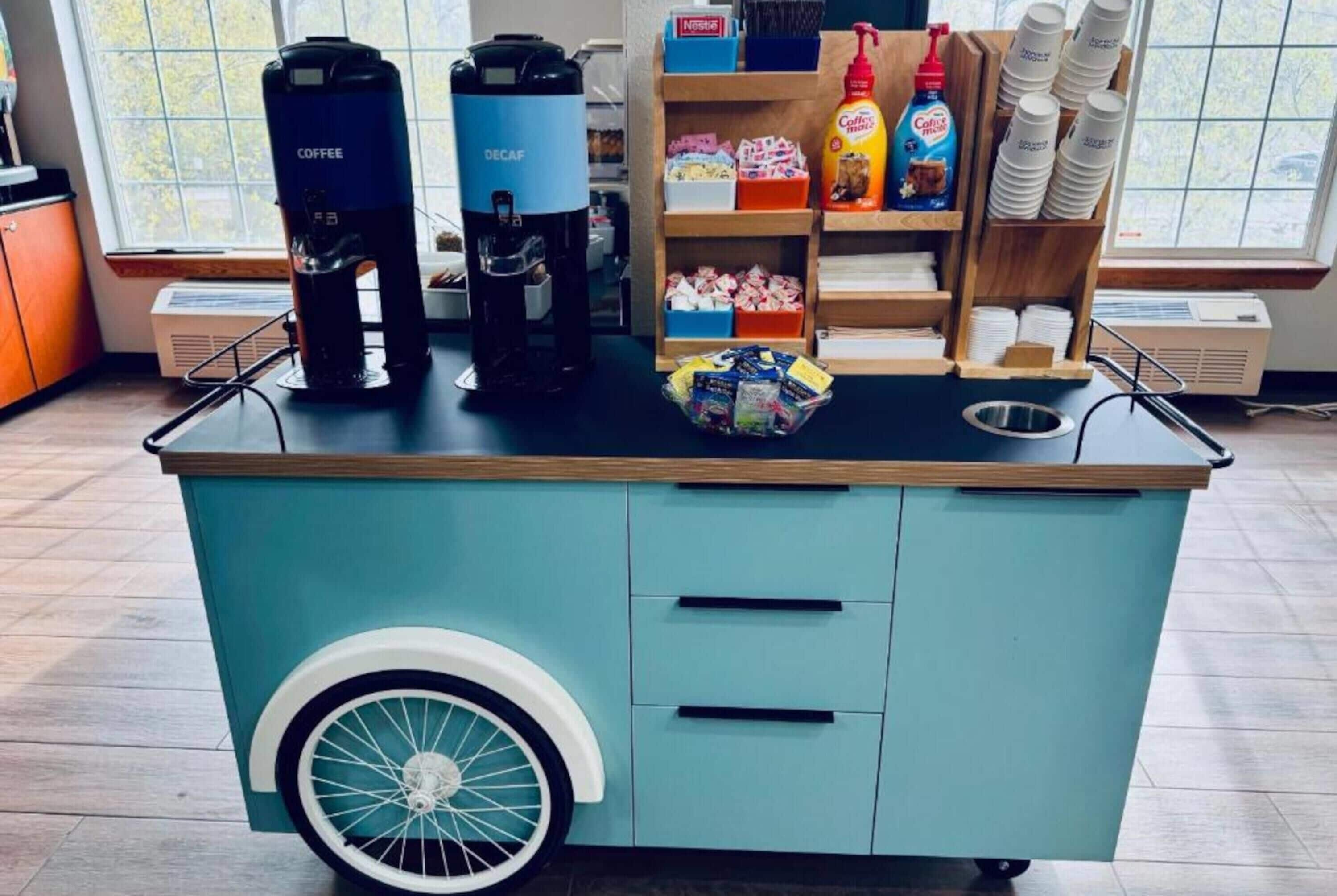 Een koffiewagen met twee koffieautomaten, een plank met koffiekopjes en toevoegingen, en een mand met koffiecups.