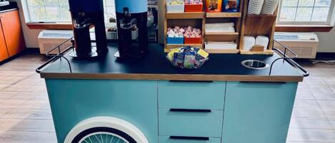 Un carrito de café con dos máquinas de café, una estantería con tazas y complementos, y una cesta de cápsulas de café.