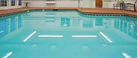 Una piscina interior con carriles delimitados, un borde de azulejos y una pequeña zona de estar con mesas y sillas.