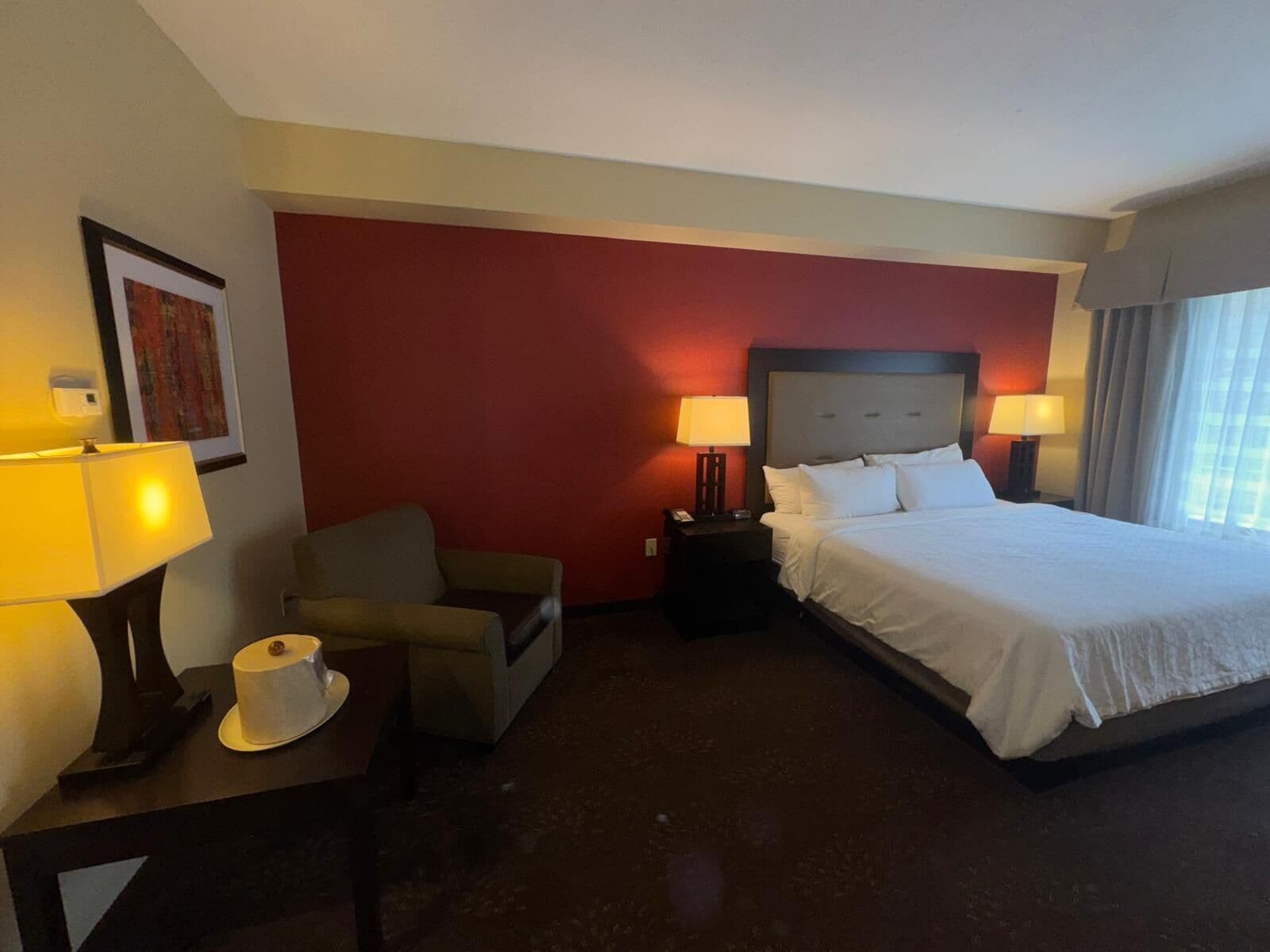 A hotel room with a bed, two bedside tables, a chair, and a desk with a lamp and a cup.