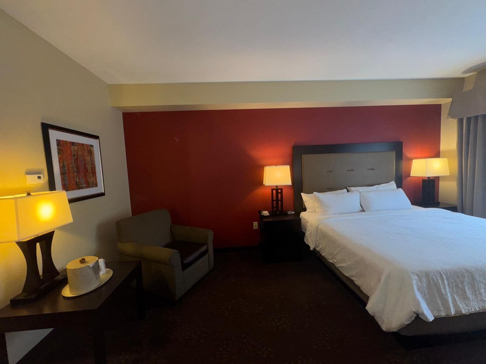 A hotel room with a large bed, two bedside lamps, a chair, a small table with a cup, and a framed picture on the wall.