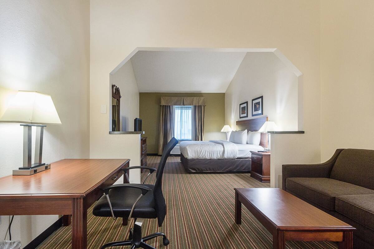 A hotel room with a wooden desk, a black office chair, a bed with a headboard, a brown sofa, and a nightstand with a lamp.