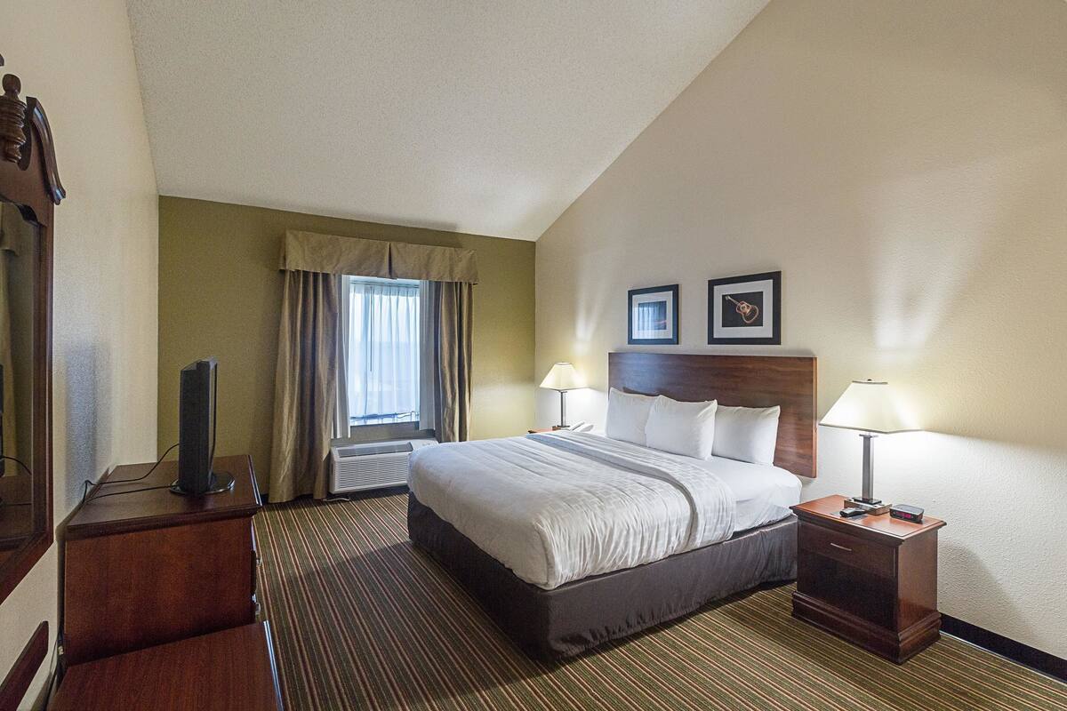 A hotel room with a large bed, two bedside lamps, a flat-screen TV, a wooden nightstand, and a window with curtains.