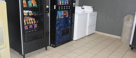 A vending machine with snacks and drinks, a small refrigerator, and a trash can in a room with tiled flooring.