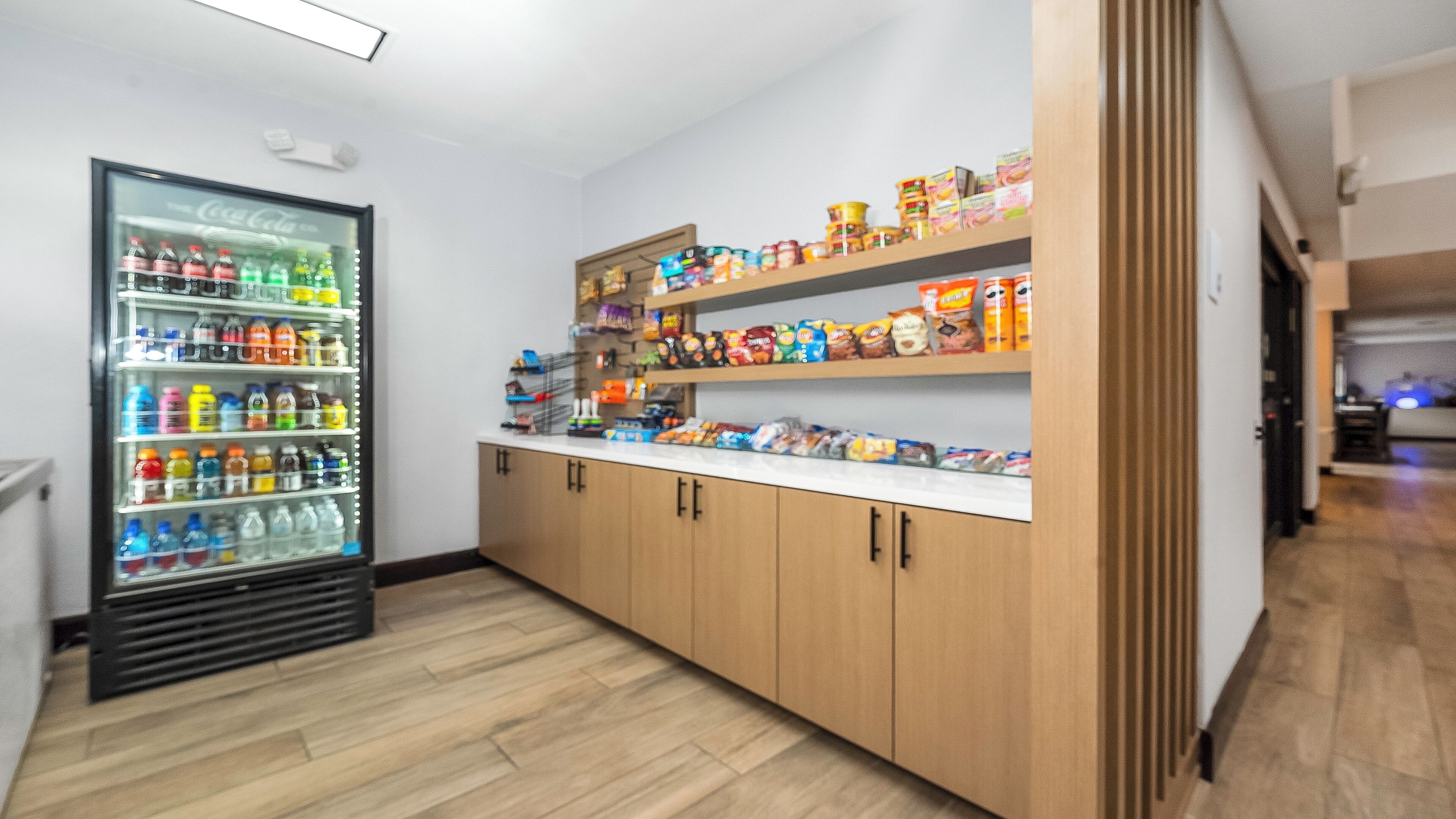 A kitchenette with a refrigerator stocked with beverages, shelves with snacks, and wooden cabinets.