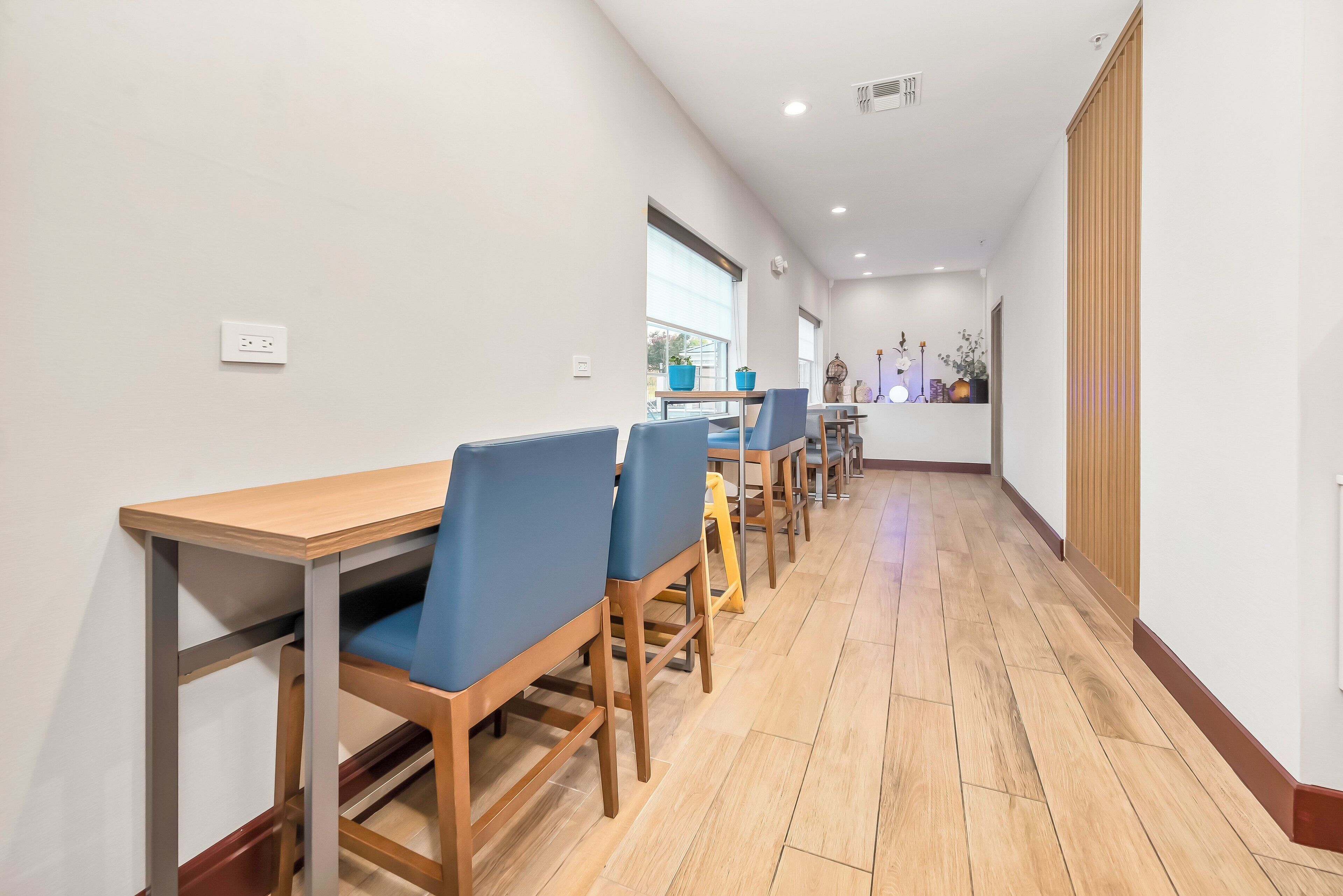 A dining area with wooden tables and blue chairs, a wooden floor, and a view of the outside through a window.