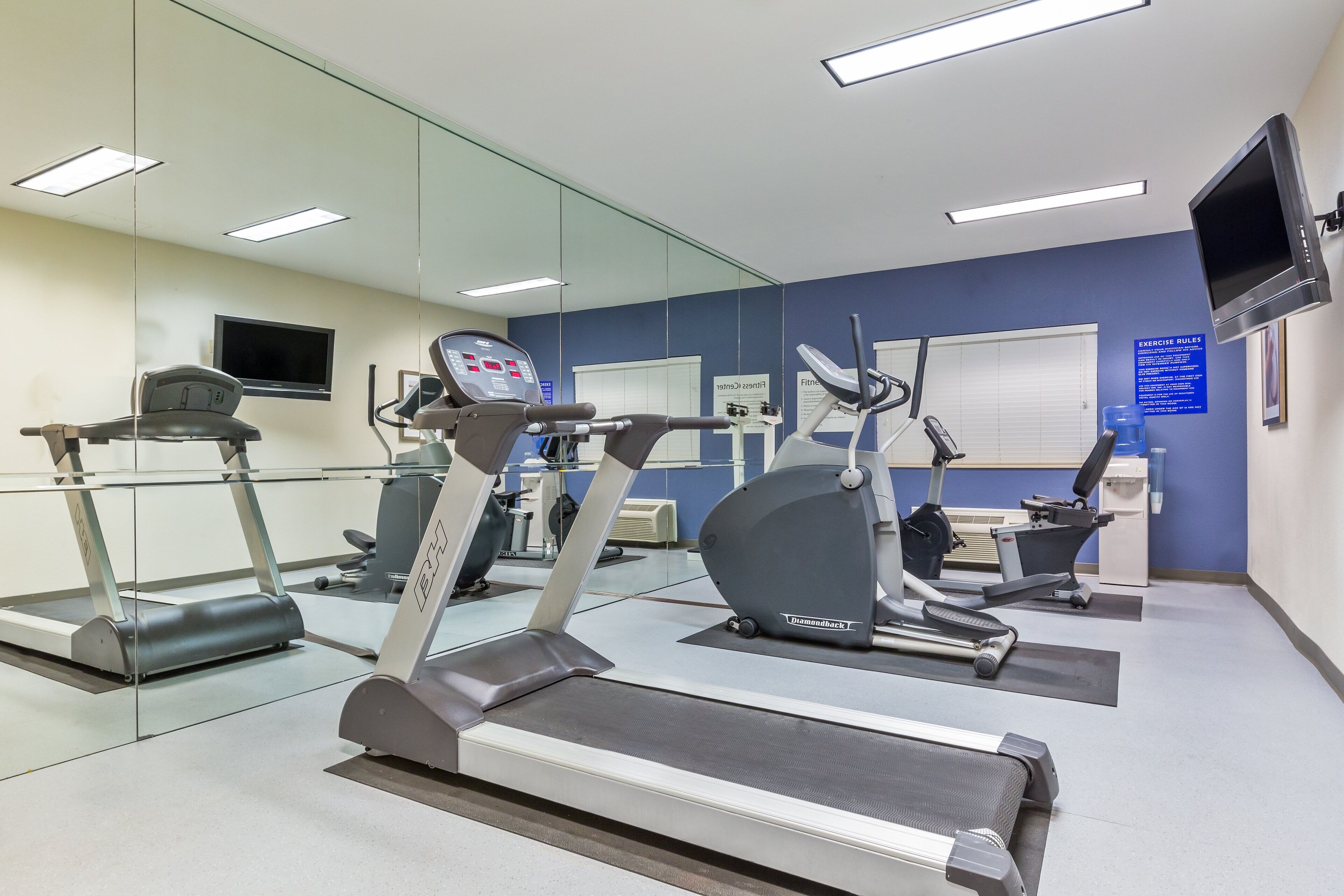 Un gimnasio con varias máquinas de ejercicio, una televisión y un dispensador de agua.