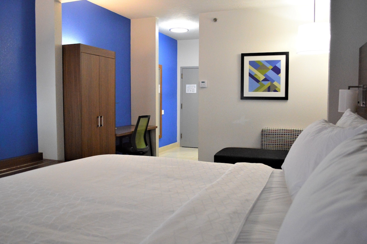 A hotel room with a bed, a desk, a chair, a wardrobe, and a wall-mounted artwork.