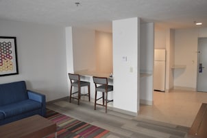 Una sala de estar con un sofá azul, una mesa de centro y un tapete a rayas de colores. Hay un pequeño comedor con una mesa y tres sillas. Se ve un refrigerador al fondo.