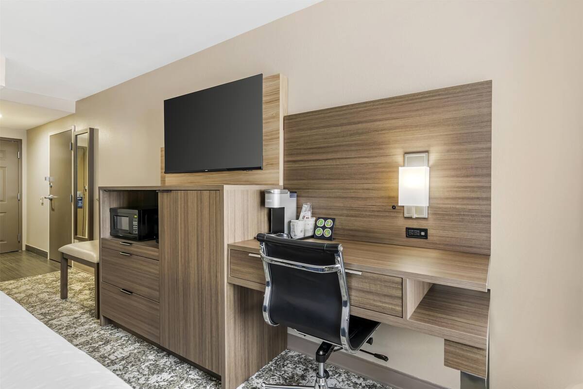 A hotel room with a wooden desk, a flat-screen TV, a microwave, a chair, and a bedside table.