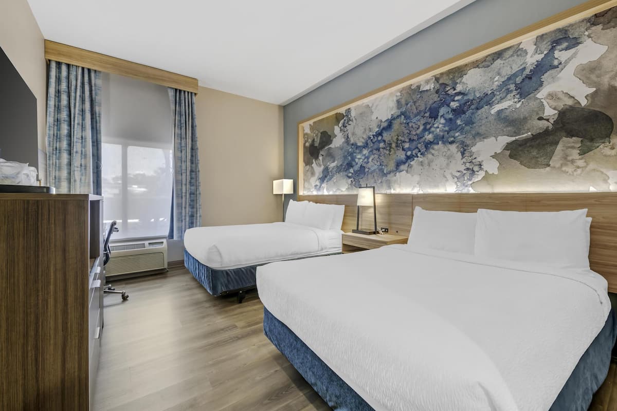 A hotel room with two beds, a large abstract painting on the wall, a desk with a lamp, and a view of the outdoors through the window.