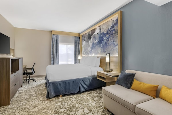 A hotel room with a bed, a desk, a chair, a sofa, a TV, and a large abstract painting on the wall.