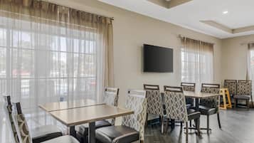 A dining area with tables and chairs, a flat-screen TV mounted on the wall, and large windows with sheer curtains.