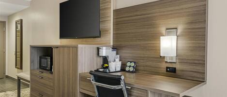 A hotel room with a wooden desk, a black chair, a flat-screen TV, a coffee machine, and a small table with cups and a tray.
