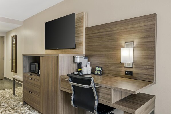 A hotel room with a wooden desk, a black chair, a flat-screen TV, a coffee machine, and a small table with cups and a tray.