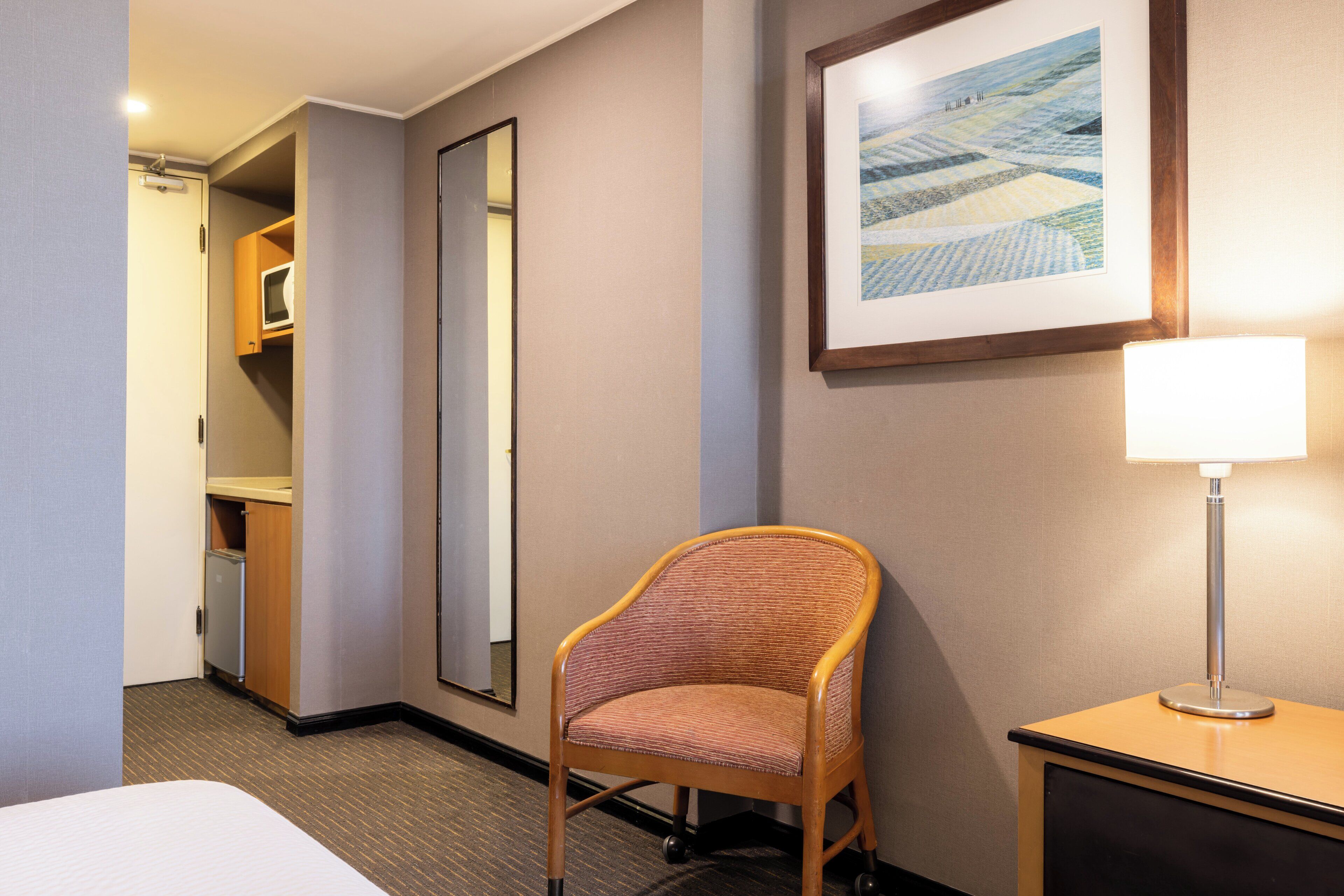 Habitación de hotel con cama, una silla, escritorio con lámpara, espejo y un cuadro en la pared.