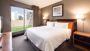 Habitación de hotel con una cama grande, un escritorio, una silla, una mesita de noche y vistas a la ciudad a través de una puerta corrediza.