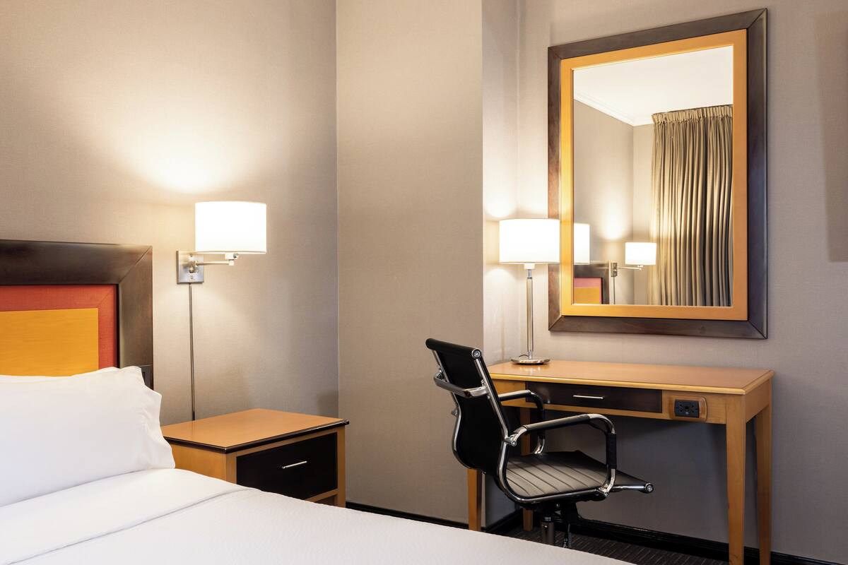 A hotel room with a bed, a desk, a chair, a nightstand, and a large mirror.