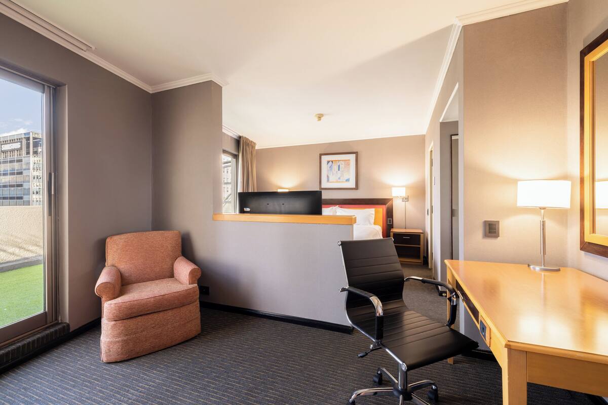 A hotel room with a large window, a desk, a chair, a bed, and a bedside table with a lamp.