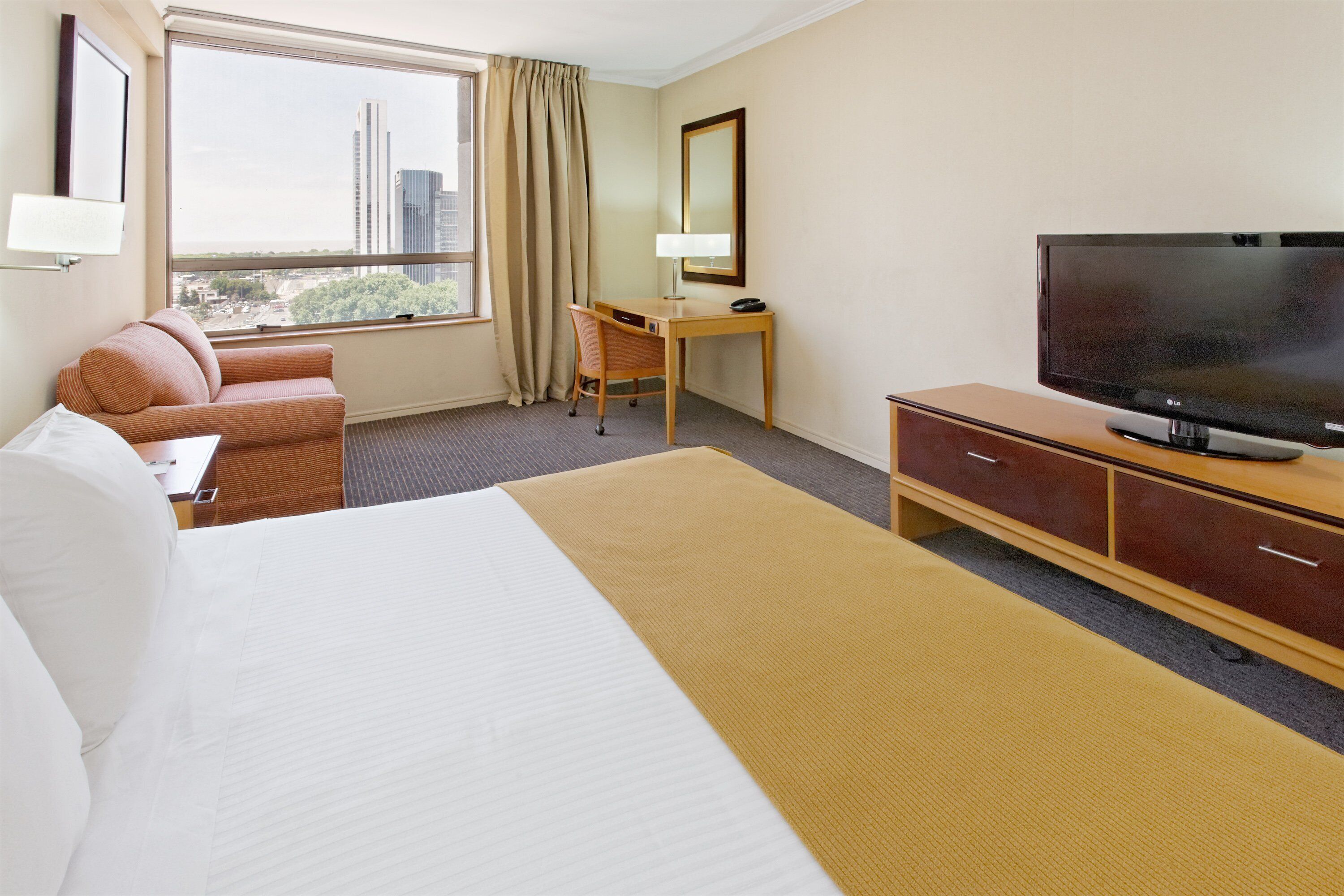 A hotel room with a bed, a desk, a television, and a view of the city.