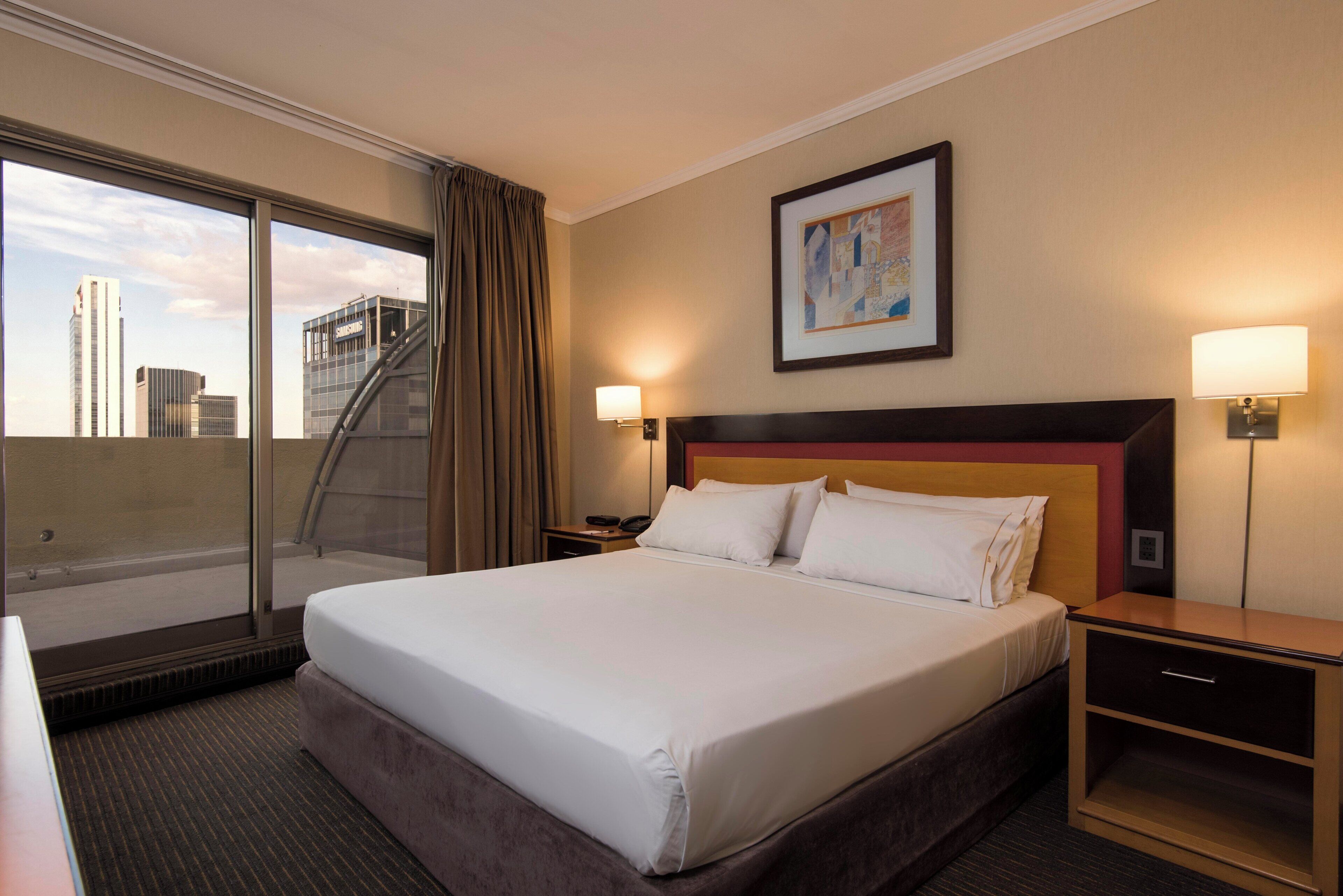 Habitación de hotel con una cama grande, un escritorio, una silla, un ventanal con vistas a edificios, un cuadro en la pared y dos lámparas de noche.