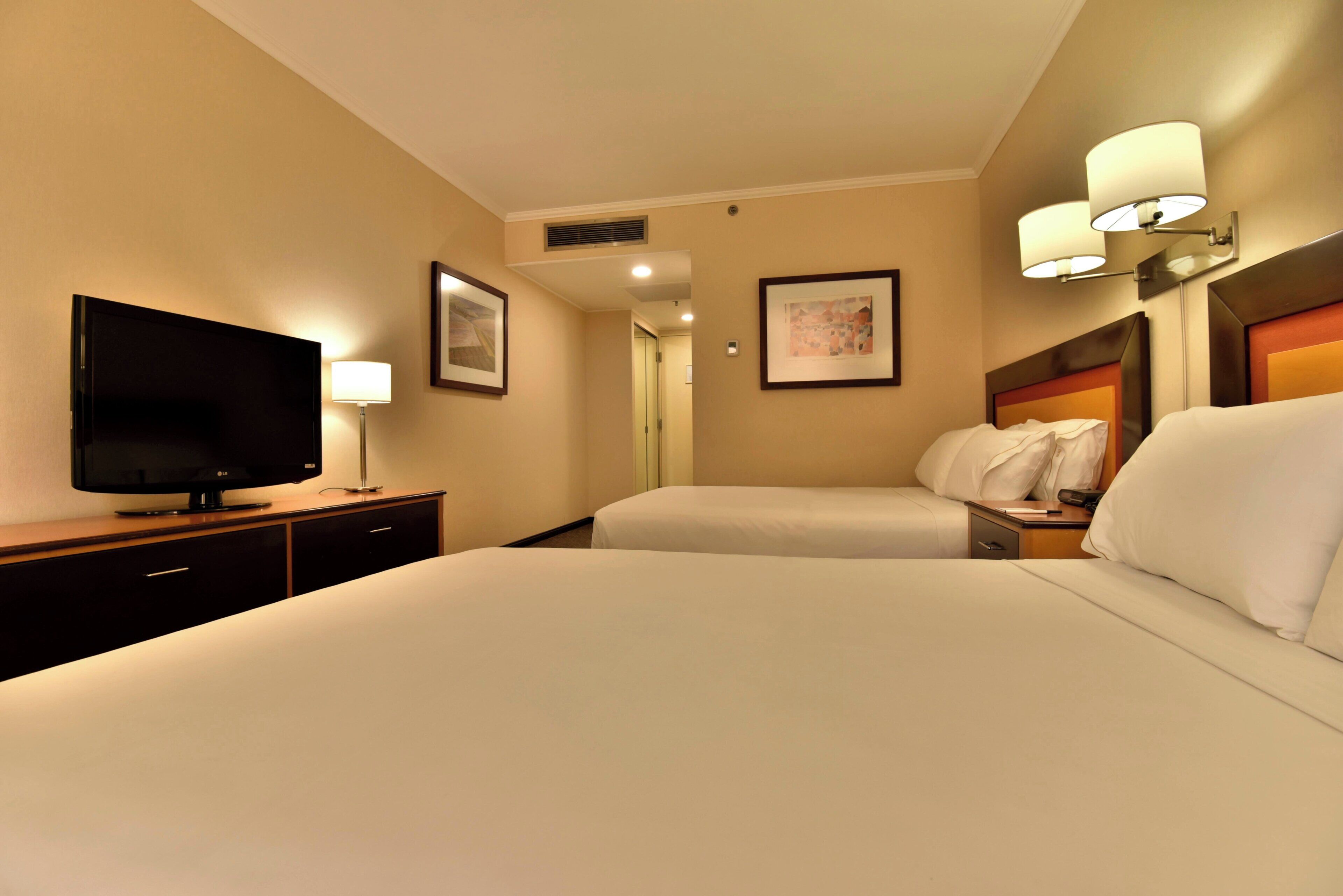 A hotel room with a flat-screen TV, a bedside table with a lamp, two beds, and a framed picture on the wall.