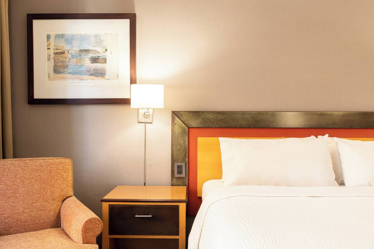 A hotel room with a bed, a nightstand, a lamp, a framed picture on the wall, and a chair.