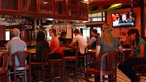 Un espace bar avec des chaises et des tables en bois, un comptoir de bar orné de bouteilles et de verres, et un écran de télévision diffusant un événement sportif.