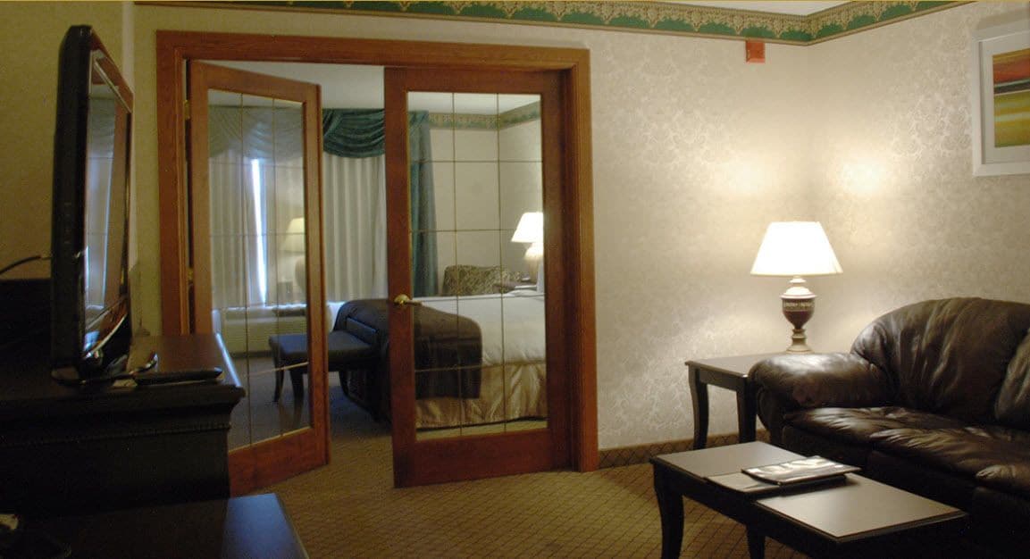 A hotel room with a leather sofa, a small table, a lamp, and a mirror. There is a door leading to a bedroom with a bed and curtains.