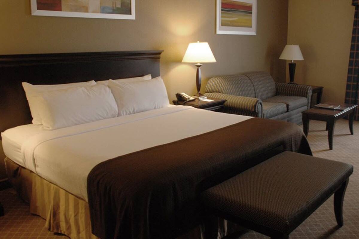 Une chambre d’hôtel avec un grand lit, une table de chevet avec une lampe, une chaise, une petite table et une fenêtre avec des rideaux.