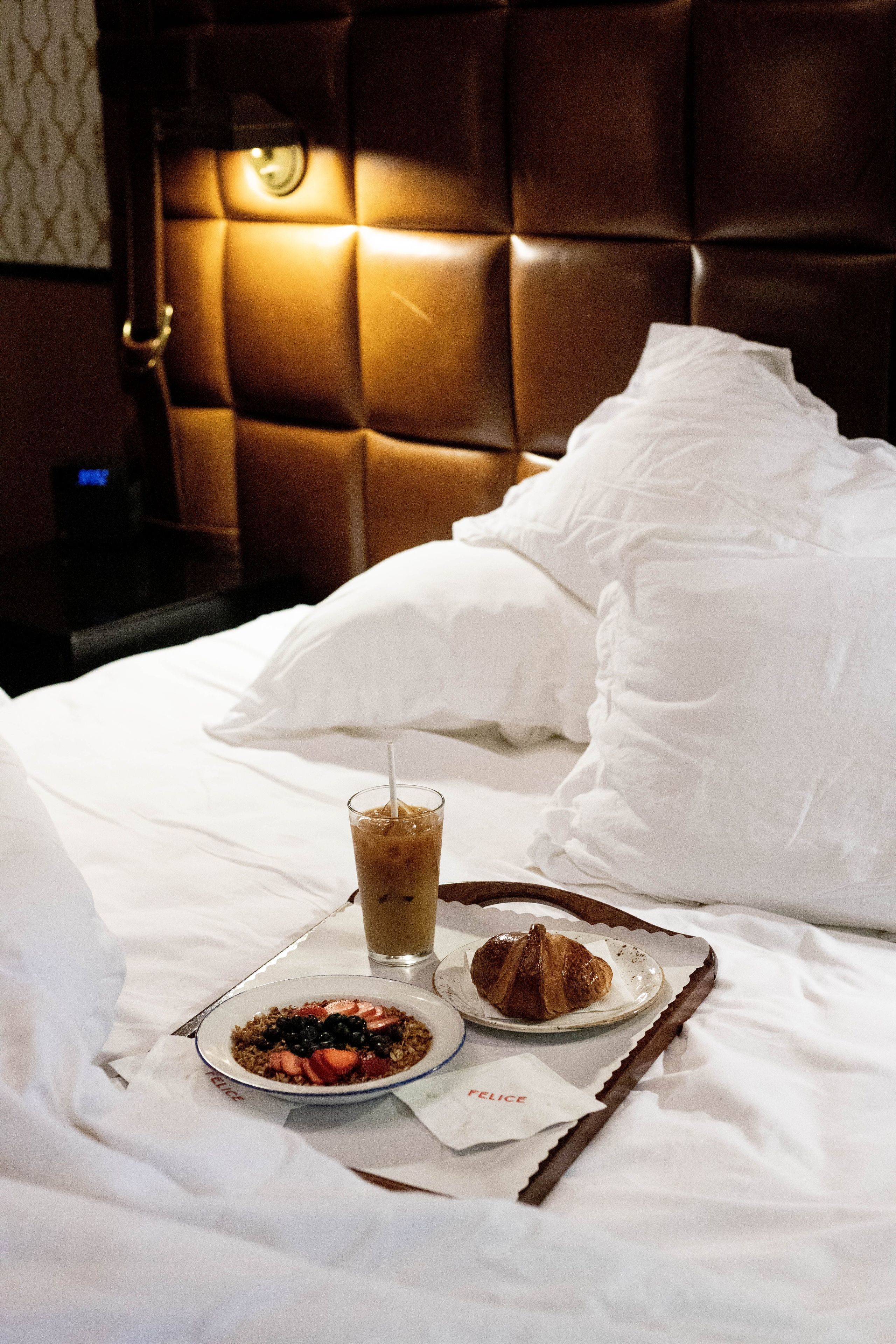 A neatly made bed with white linens, a tray with breakfast items including a croissant, a glass of iced coffee, and a bowl of cereal.