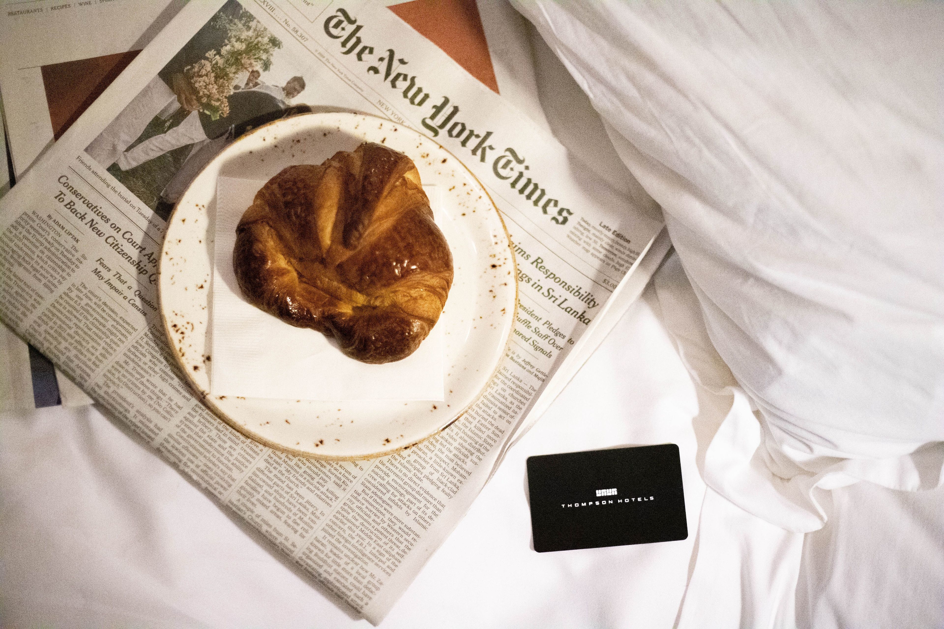 A plate with a pastry on a newspaper, a black card, and white bedding.