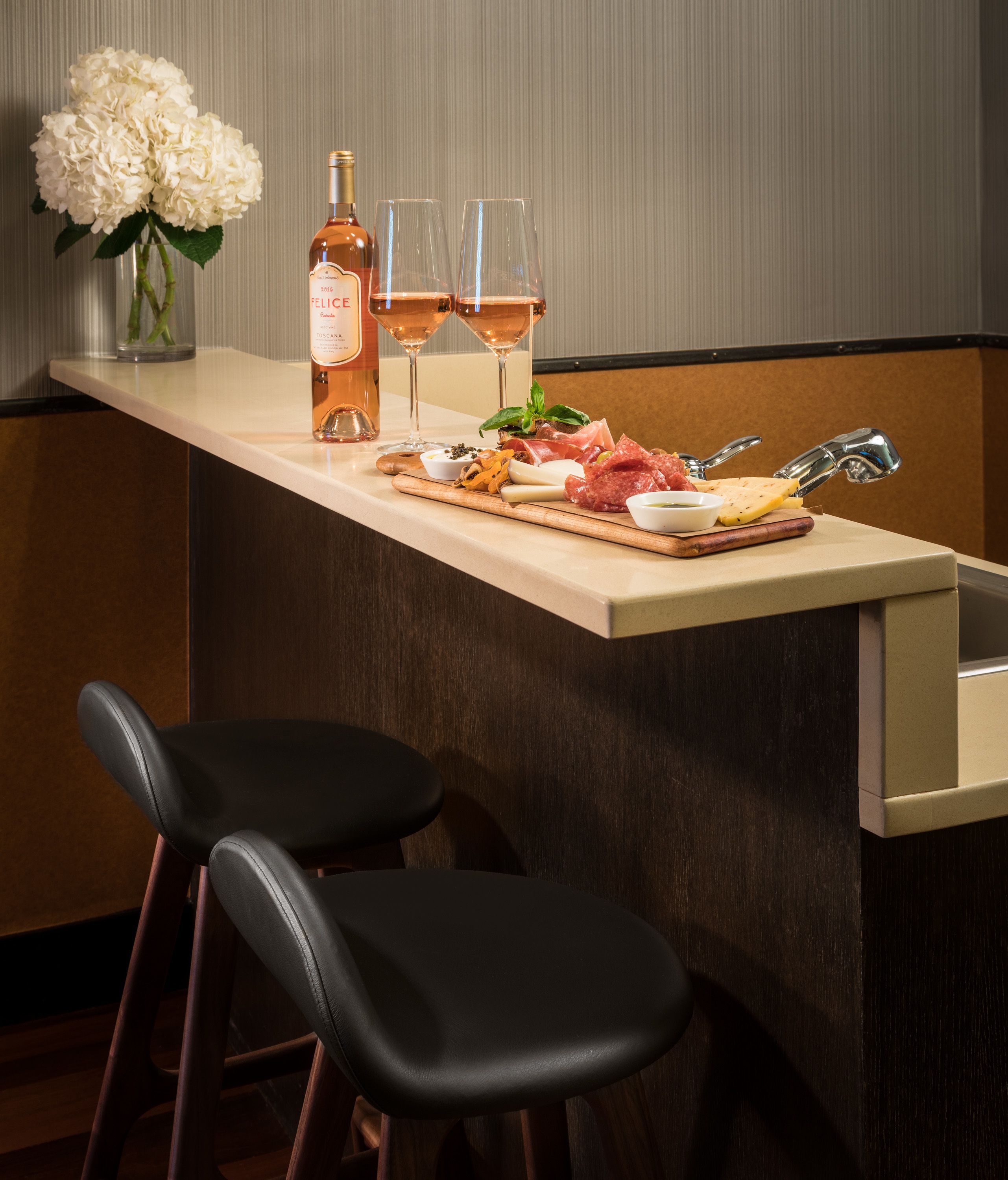 A bar area with a bottle of wine, glasses, a cheese board, and a vase of flowers.
