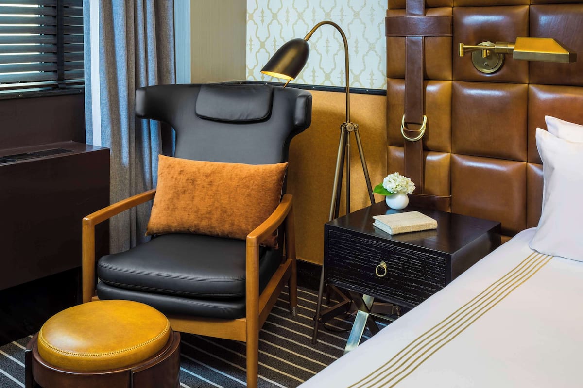 Une chambre d’hôtel avec un fauteuil en cuir, une table de chevet avec une lampe, un petit vase avec des fleurs et un lit soigneusement fait.