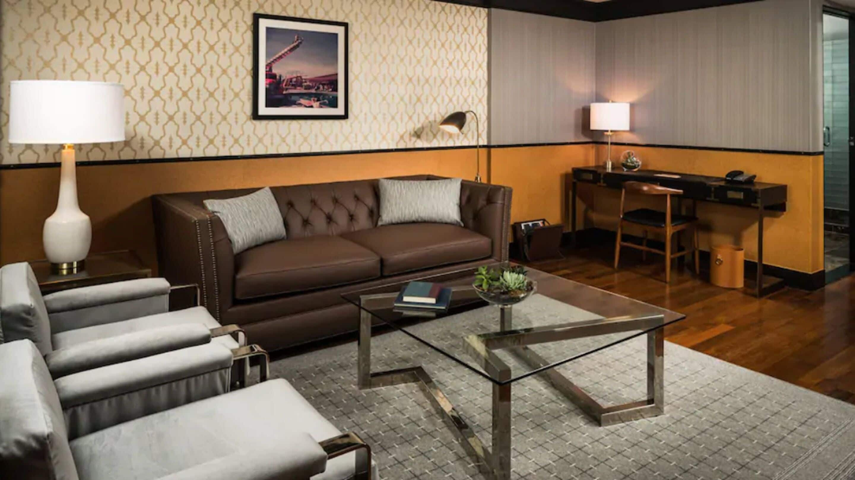 A hotel room with a brown leather sofa, a glass coffee table, and a grey patterned rug. There is a desk with a chair and a lamp.