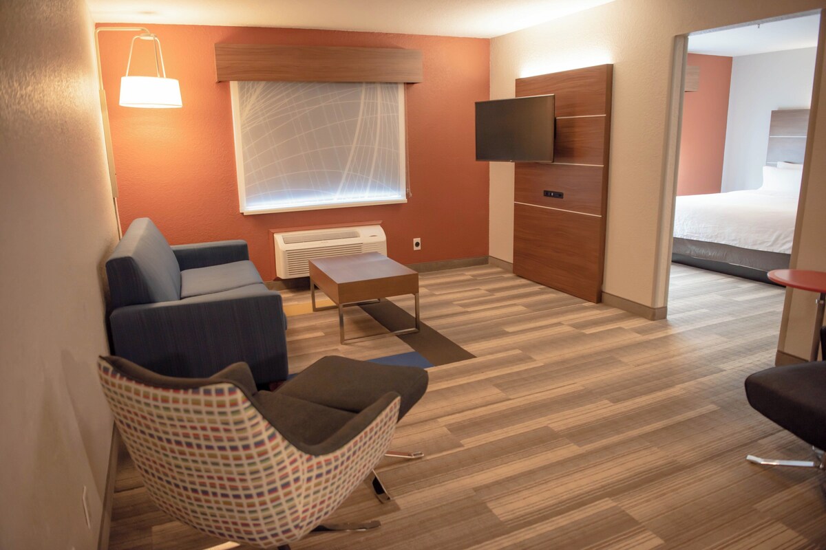 A modern hotel room with a sofa, a patterned chair, a small table, a flat-screen TV mounted on the wall, and a bed in the adjacent room.
