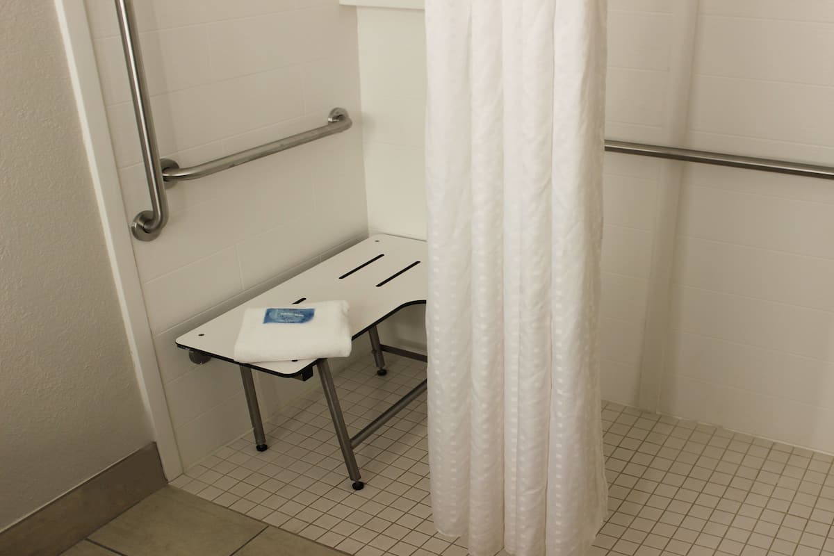 A bathroom with a white shower curtain, a white towel on a metal shelf, and a handrail.