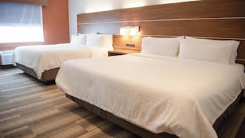 A hotel room with two beds, a wooden headboard, a nightstand with a lamp, and a window with blinds.