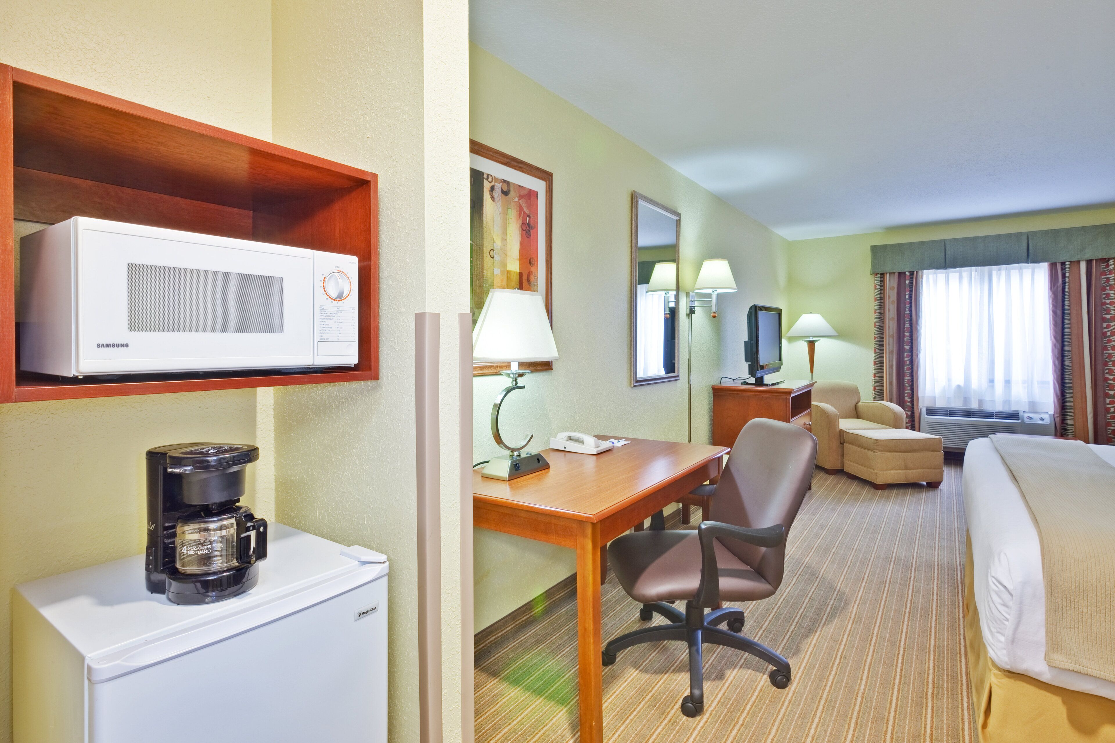 A hotel room with a bed, a desk with a chair, a microwave, a coffee maker, a lamp, and a television.
