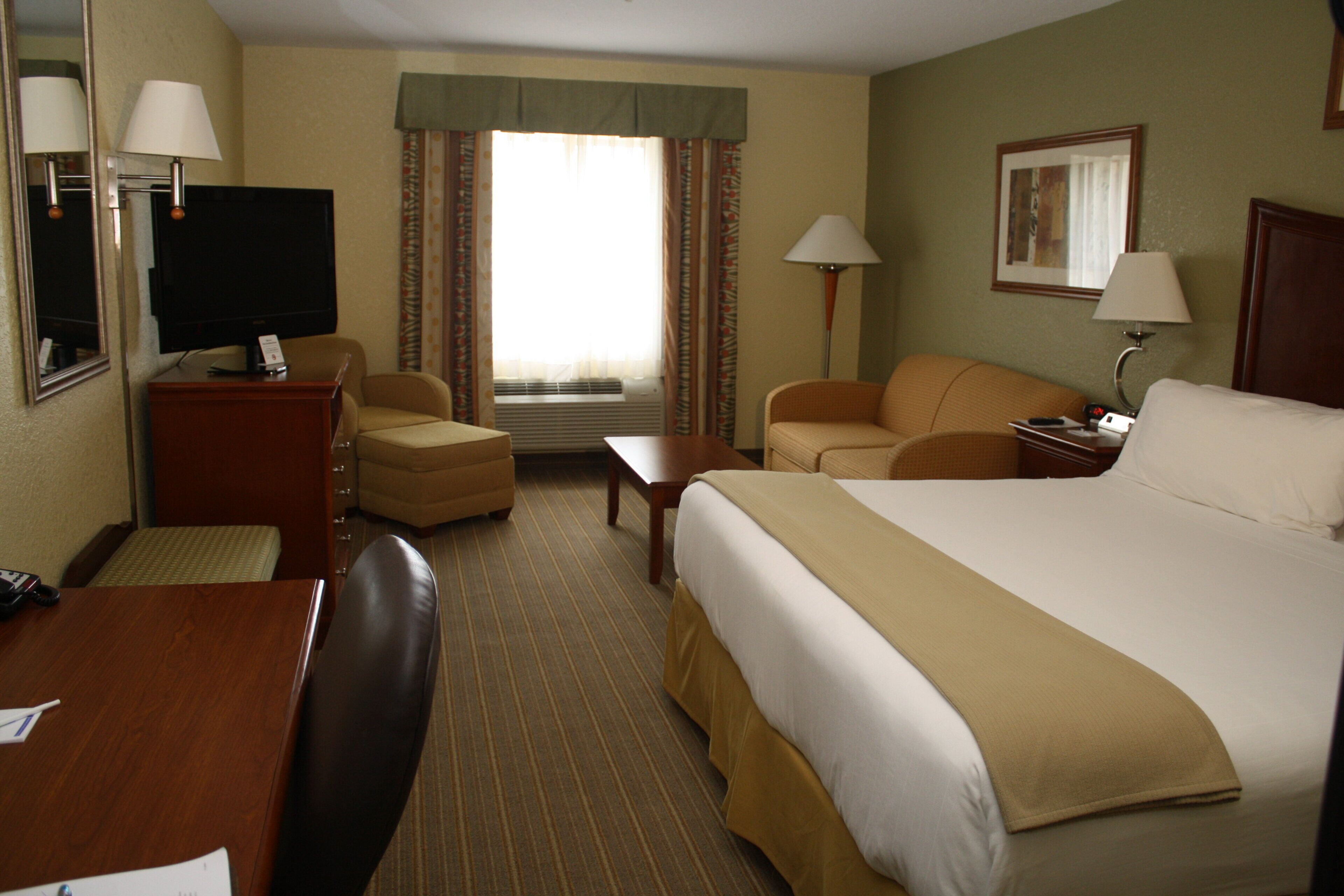 A hotel room with a large bed, two armchairs, a desk with a television, and a window with curtains.