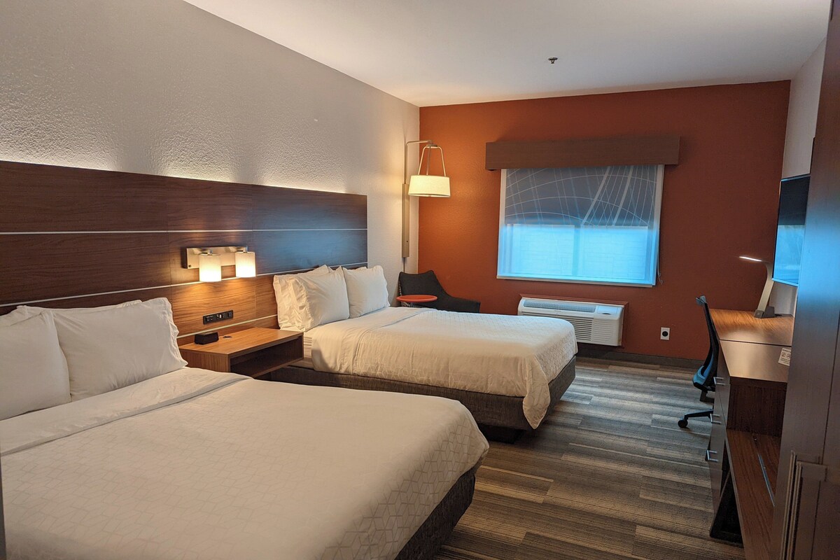 A hotel room with two beds, a desk, a chair, a lamp, and a window.