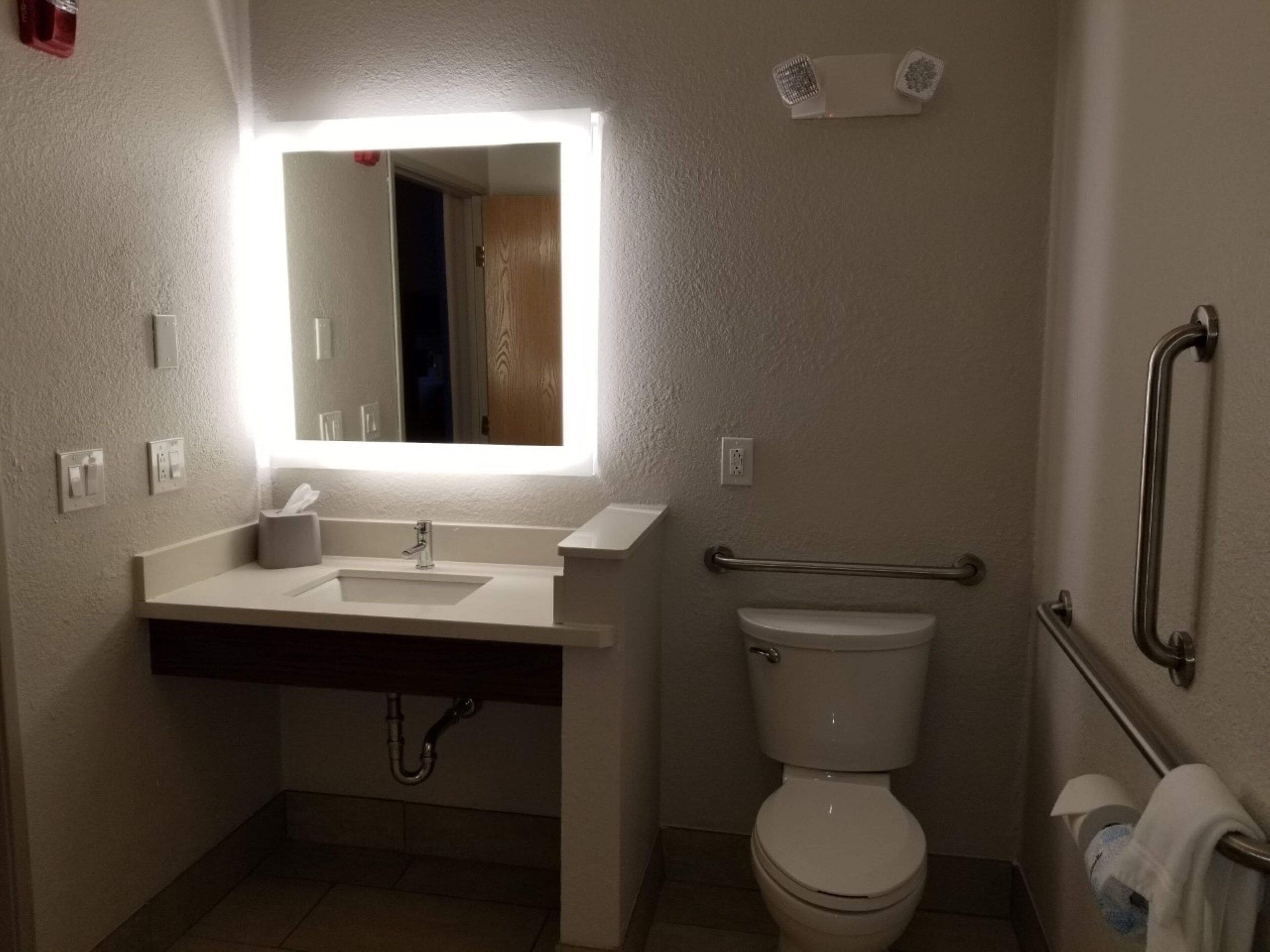A bathroom with a white sink, mirror, and toilet. There is a handrail on the right side and a towel rack on the left.