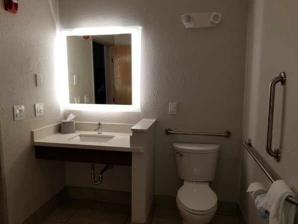 A bathroom with a white sink, mirror, and toilet. There is a handrail on the right side and a towel rack on the left.
