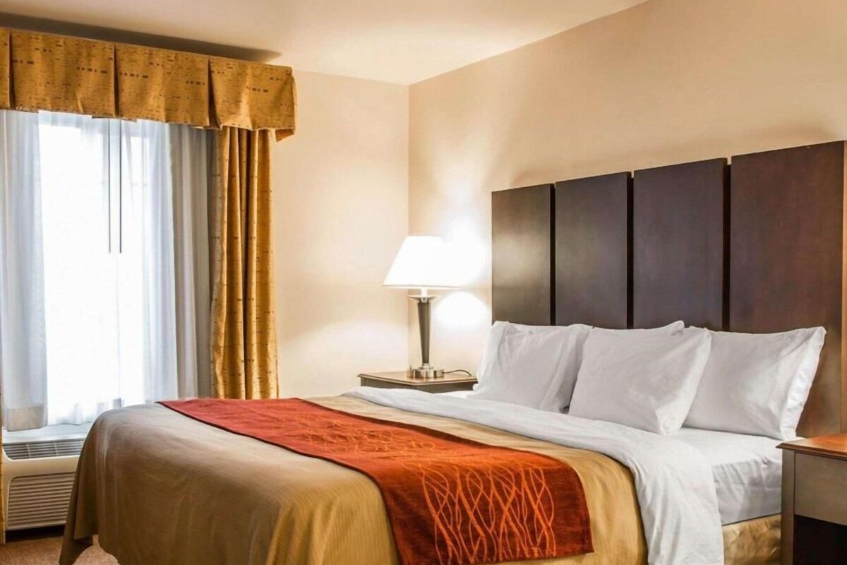 Habitación de hotel con cama, mesita de noche con lámpara, ventana con cortinas y cabecero fijado a la pared.